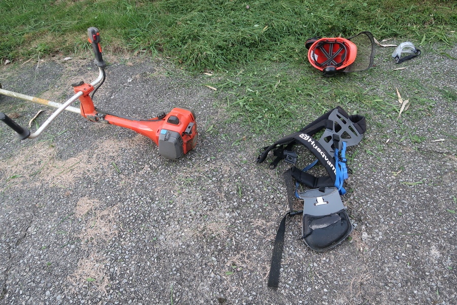 orange strimmer, chest harness, helmet and goggles strewn on a concrete slab